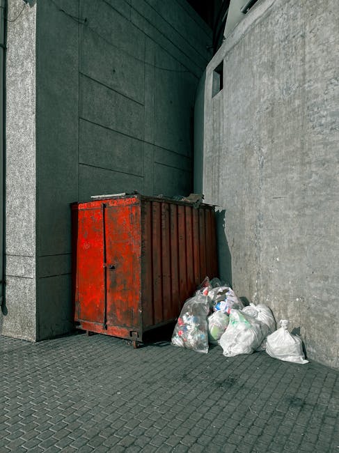 A large, red metal rubbish dumpster with a weathered surface and visible rust patches, situated on a paved outdoor area against a dark green wall and a light gray concrete wall. The dumpster is positioned at an angle, with its door facing slightly toward the viewer, and is partially open with some waste inside. Surrounding the base of the dumpster, there are several clear plastic bags filled with generic rubbish, including plastic bottles and packaging, placed on the textured paving slabs. The area appears to be a designated waste disposal point in an urban setting, possibly part of a private waste collection service such as Waste Collection Bromley, which offers alternative rubbish removal options. The environment is clean yet utilitarian, with minimal environmental context visible, just the surrounding walls and pavement, highlighting the focused scene of waste ready for collection or disposal as part of an independent rubbish clearance solution.