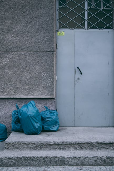 A large, red metal rubbish dumpster with a weathered surface and visible rust patches, situated on a paved outdoor area against a dark green wall and a light gray concrete wall. The dumpster is positioned at an angle, with its door facing slightly toward the viewer, and is partially open with some waste inside. Surrounding the base of the dumpster, there are several clear plastic bags filled with generic rubbish, including plastic bottles and packaging, placed on the textured paving slabs. The area appears to be a designated waste disposal point in an urban setting, possibly part of a private waste collection service such as Waste Collection Bromley, which offers alternative rubbish removal options. The environment is clean yet utilitarian, with minimal environmental context visible, just the surrounding walls and pavement, highlighting the focused scene of waste ready for collection or disposal as part of an independent rubbish clearance solution.