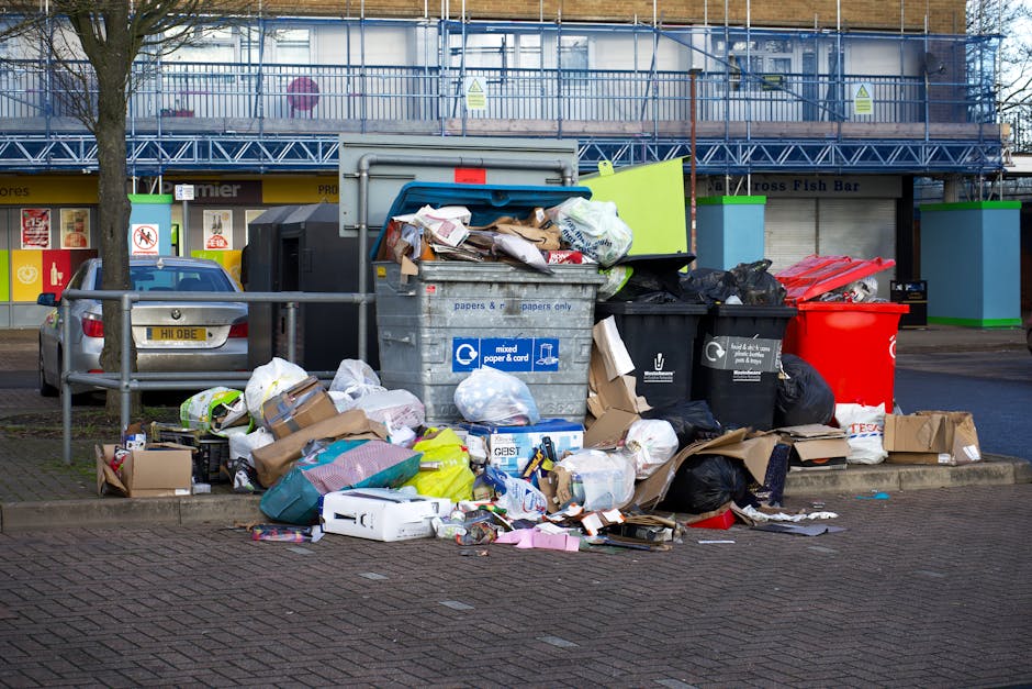 A large collection of overflowing rubbish and recycling bins situated on a paved sidewalk area in front of a commercial building. The collection includes a grey mixed paper and card bin filled with flattened cardboard boxes, paper, and packaging materials, with additional waste such as plastic bags, cardboard boxes, and discarded packaging scattered around its base. To the right, there are black and red trash bins, some with rubbish spilling over and others closed. Behind the bins, a metal railing separates the waste area from the adjacent street, where a parked silver car is partially visible. The background features a building with a blue facade, yellow signage, and a covered outdoor area with a blue scaffold or construction scaffolding present, indicating ongoing work. The scene is lit with natural daylight, emphasizing the cluttered and unmanaged state of the waste collection, illustrating an instance of private rubbish disposal that Waste Collection Bromley might address through alternative waste handling services.