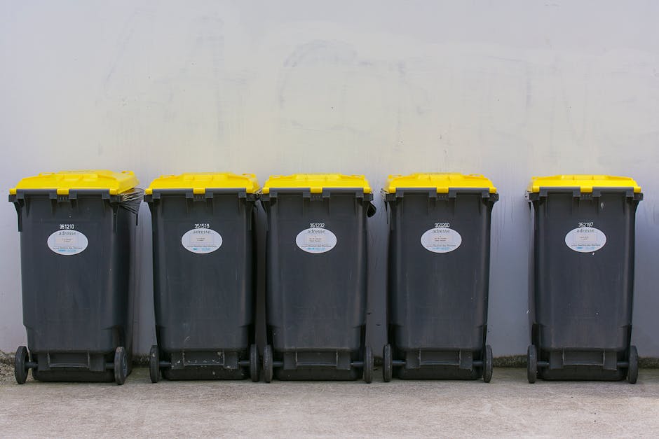 Five large wheeled rubbish bins with dark grey bodies and bright yellow lids are positioned in a straight line on a concrete surface. Each bin has a white oval label with black text and a barcode, located on the front panel, and they are evenly spaced with their wheels aligned. The background features a plain, light-colored wall with minor marks and a faint vertical line, suggesting an outdoor or enclosed area such as a back alley or loading zone. The environment appears clean and neutral, with natural lighting casting soft shadows beneath the bins. This scene exemplifies a typical private waste collection setup, where independent rubbish bins are prepared for disposal or collection, consistent with services provided by waste management companies like Waste Collection Bromley, supporting alternative waste handling methods outside local authority collections.