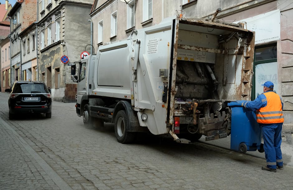 A large collection of overflowing rubbish and recycling bins situated on a paved sidewalk area in front of a commercial building. The collection includes a grey mixed paper and card bin filled with flattened cardboard boxes, paper, and packaging materials, with additional waste such as plastic bags, cardboard boxes, and discarded packaging scattered around its base. To the right, there are black and red trash bins, some with rubbish spilling over and others closed. Behind the bins, a metal railing separates the waste area from the adjacent street, where a parked silver car is partially visible. The background features a building with a blue facade, yellow signage, and a covered outdoor area with a blue scaffold or construction scaffolding present, indicating ongoing work. The scene is lit with natural daylight, emphasizing the cluttered and unmanaged state of the waste collection, illustrating an instance of private rubbish disposal that Waste Collection Bromley might address through alternative waste handling services.